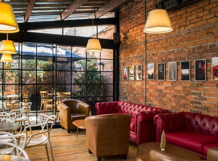 An interior featuring an exposed brick wall, glass panel ceiling, leather sofas, and tables, creating a warm ambiance.