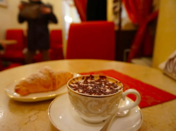 A chocolate-covered beverage served in a beautifully designed teacup, accompanied by a small plate with a croissant and a sauce.