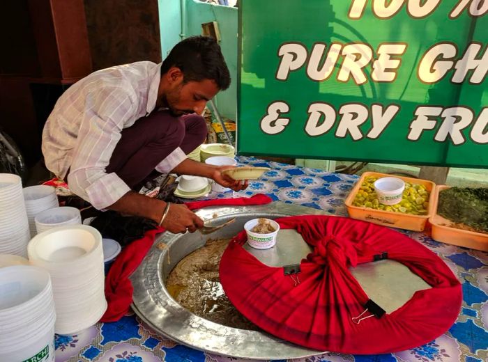 A cook crouches by a sunken pot of haleem, carefully serving some into a customer’s bowl.