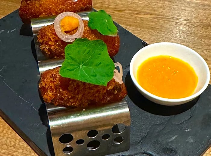 Golden fried croquettes served in a small metal holder with a dipping sauce.