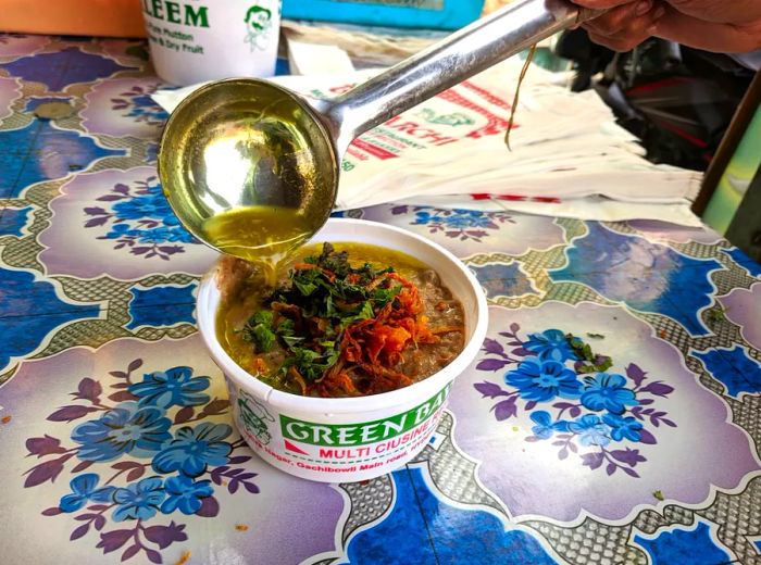 A ladle drizzles fat into a branded haleem bowl.