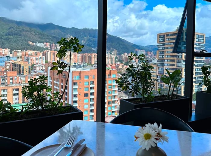 A beautifully set table with cutlery and flowers in the foreground, framed by large windows that overlook the city of Bogotá and its surrounding hills.