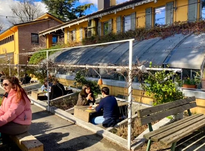 Outdoor tables arranged on different levels in a sunlit garden, where diners in winter attire savor their lunches.