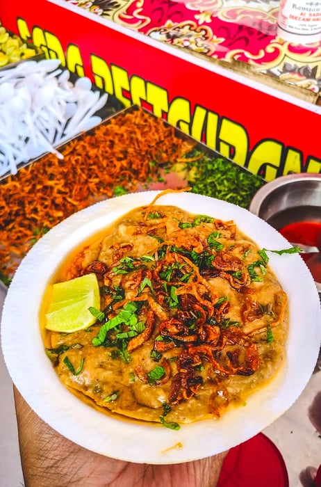 A bowl of haleem garnished with crispy fried onions, fresh herbs, and a slice of lemon.