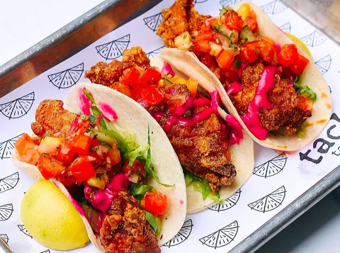 Three crispy fish tacos presented on stylish branded paper within a sheet pan.