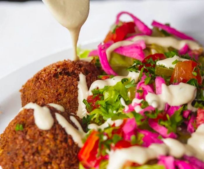 A spoon drizzles sauce over falafel served with salad.