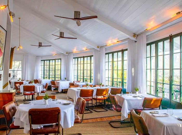 A bright dining area featuring large windows, modern chairs, and tables dressed in white tablecloths.