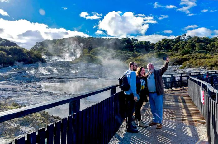 Three individuals of various ages stand on a boardwalk, posing for a selfie amidst the steam and bubbling earth behind them.