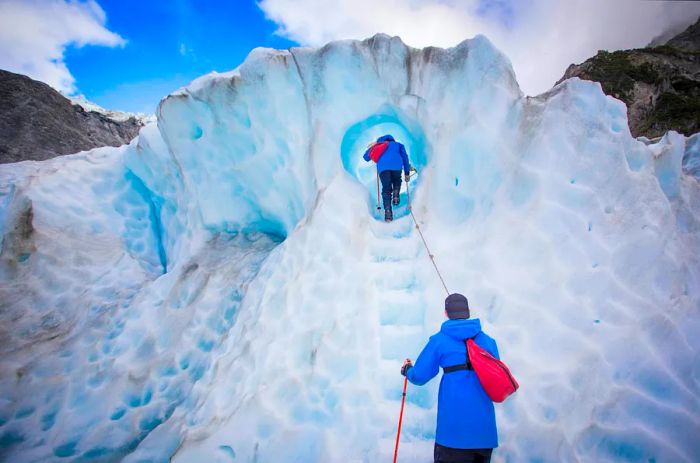 Two hikers traverse icy steps through a glacier tunnel.