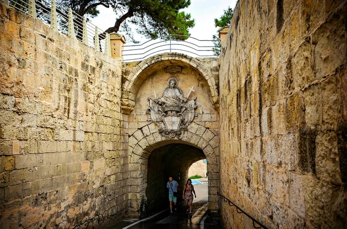 Two individuals pass through a grand stone gateway, leading into the ancient city of Mdina, Malta.