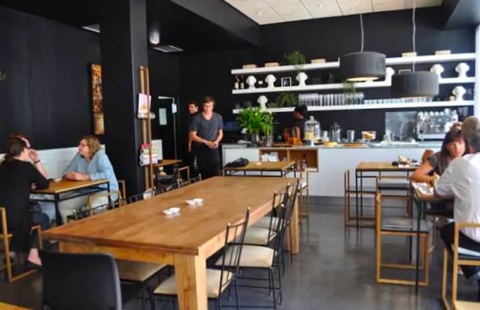 A cozy café setting filled with customers seated at small tables and a large communal table at the center, while a counter at the back showcases decorative items against the wall.