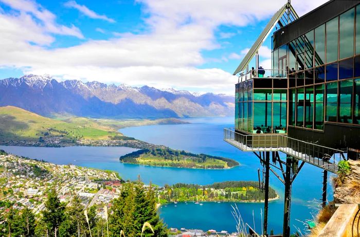 A grand glass structure perched on a hillside offers breathtaking views of the Queenstown skyline, framed by snow-covered peaks and a sprawling lake with a patch of greenery extending into the water.