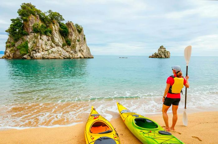 A woman gazes towards a rocky shoreline while standing beside a yellow kayak.