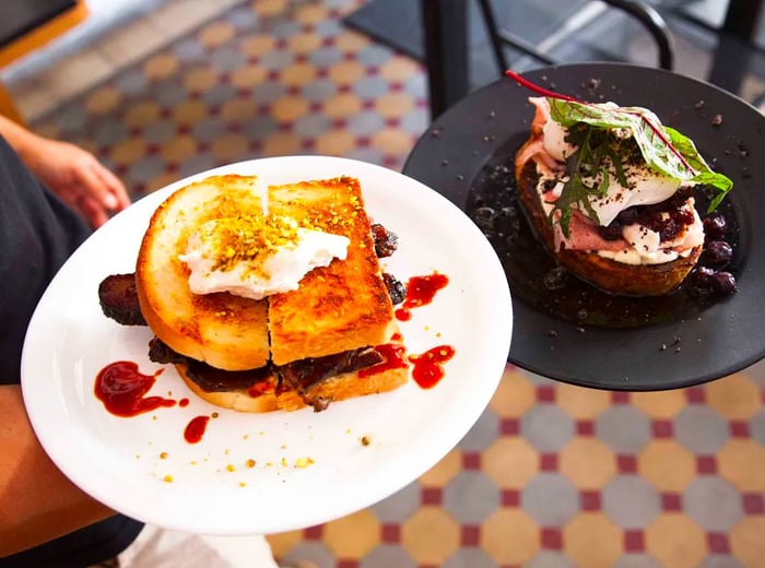 A server presents a large jam-filled sandwich topped with cream alongside a vegetable-rich dish.