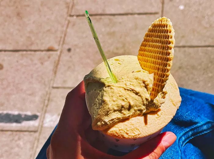 A hand holds a small cup of gelato, with a large thin waffle peeking out over the sidewalk.