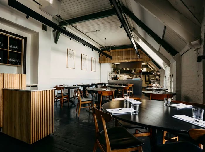 The long dining area features high wooden-beamed ceilings, simple tables arranged on a wooden floor, and an open kitchen at the far end.