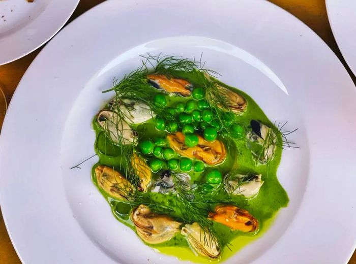 An overhead view of a bowl filled with mussels in a vibrant green sauce, garnished with peas and fresh dill.