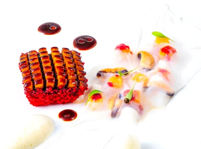 Geometrically cut squid with a vibrant red exterior is accompanied by additional squid pieces, edible flowers, and mango slices topped with a delicate white foam.