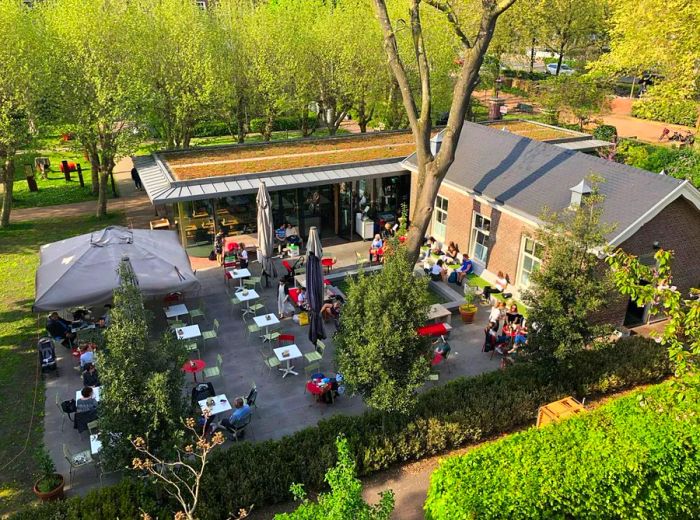 An aerial view of the outdoor patio, featuring small tables with umbrellas, bordered by hedges on one side, open greenery on another, and an L-shaped building framing the space.