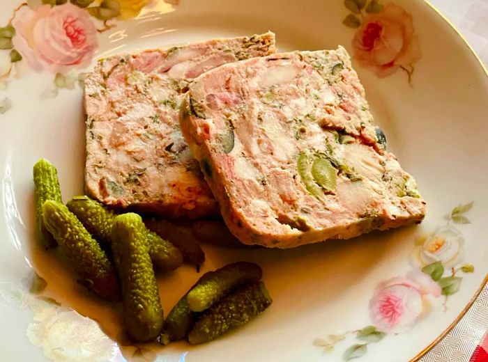 A floral-decorated plate features slices of terrine accompanied by pickles.