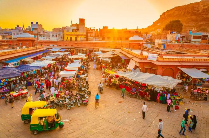 Rickshaws and motorcycles are lined up near stalls in a bustling market framed by red walls.