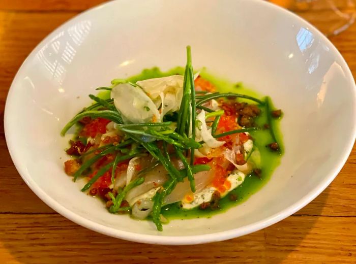 A dish featuring sliced fish in a green sauce, adorned with sprigs of samphire and flecks of orange roe.