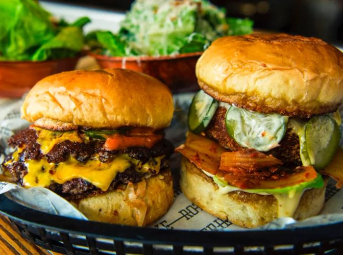 Two towering burgers, one with melted cheese and the other adorned with pickles and kimchi.