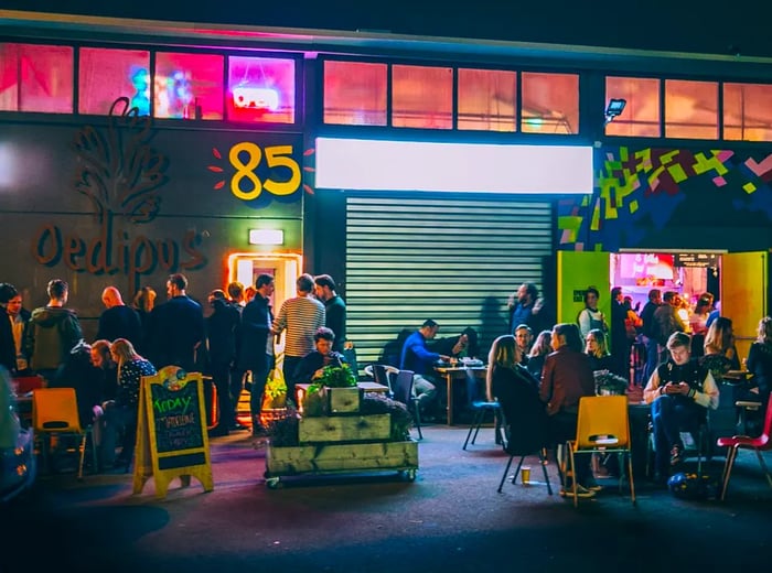 The venue features an industrial facade with neon lights illuminating the warehouse-like interior, where a lively crowd gathers outside, mingling around open doorways or relaxing on patio chairs.