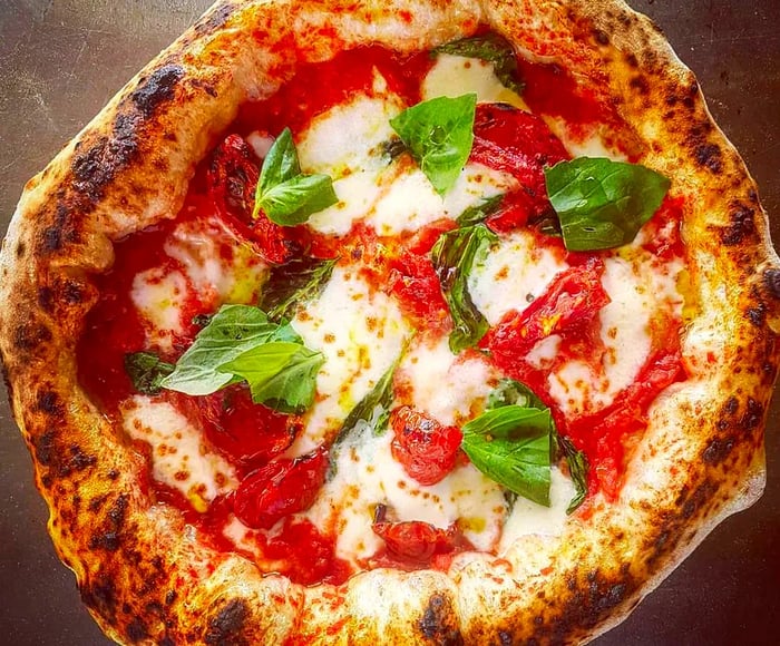 An aerial view of a charred pizza featuring a thick crust, adorned with tomato sauce, cheese, and fresh basil leaves.