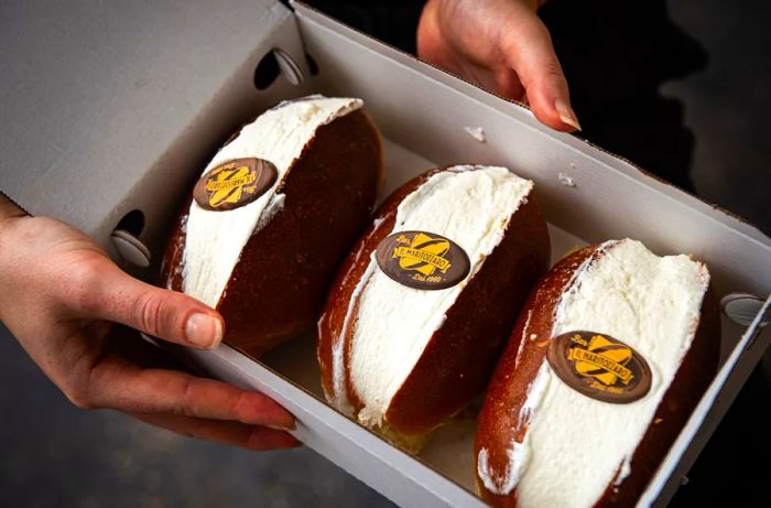 Hands grasp a box filled with cream-filled pastries adorned with edible logos for Il Maritozzaro.