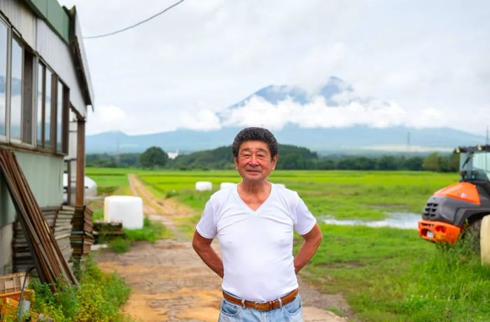 A farmer stands with his hands at his sides in front of a vast green landscape, with cloud-covered mountains rising in the background.