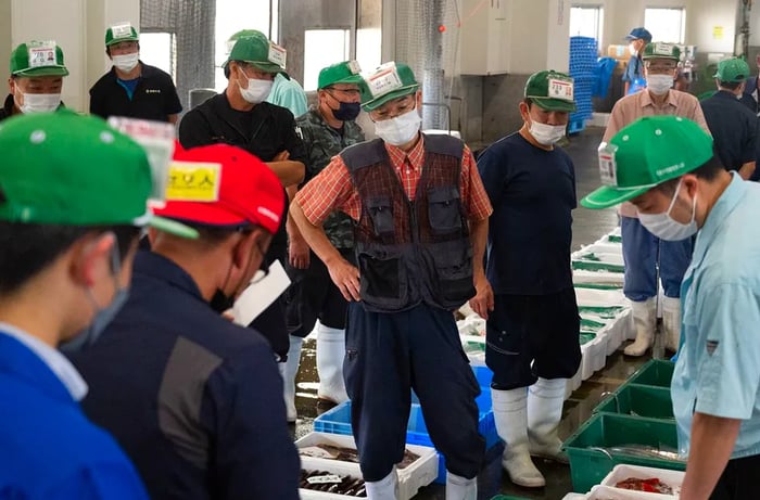 Shoppers wearing baseball caps adorned with identifying logos gather around bins filled with fresh fish for sale.