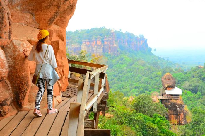 A hiker cautiously steps onto a suspended wooden walkway that hugs the mountain's edge.