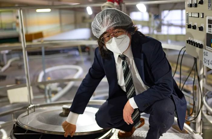 A suited man wearing a hairnet carefully opens a metal cask of sake.