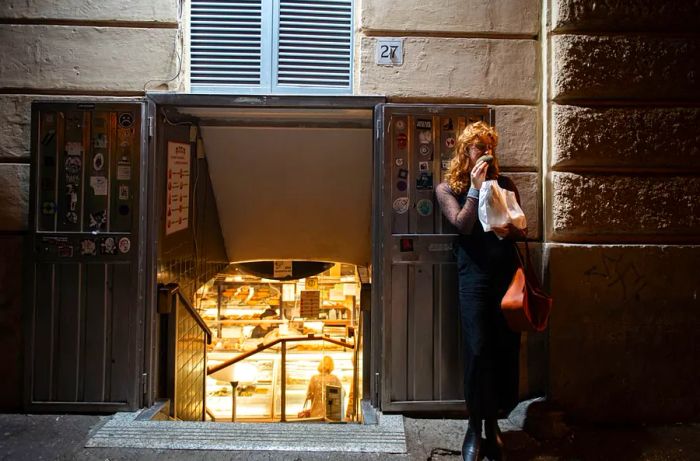 A woman enjoys a pastry while leaning against the wall of a bakery, with light spilling from the entrance down the stairway next to her.