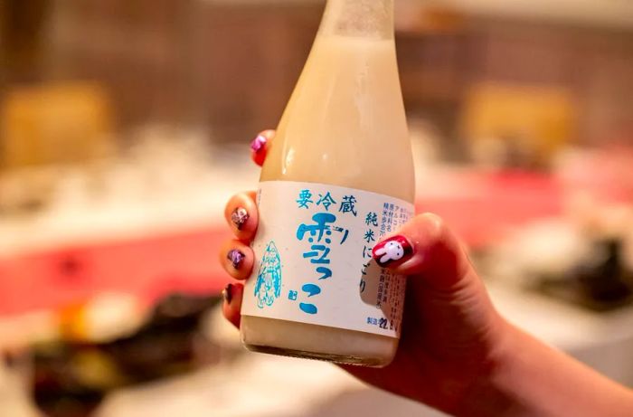 A hand with beautifully painted nails lifts a cloudy bottle of sake.