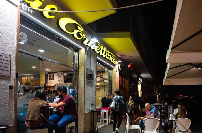 Diners enjoy their meals at tables outside a bakery under the night sky.