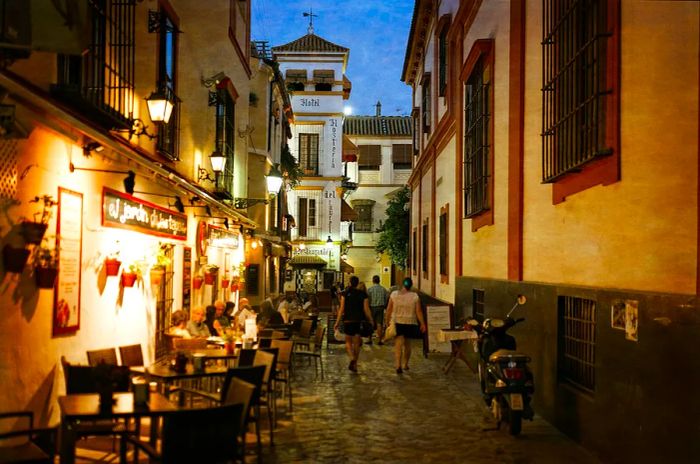 People stroll past tables on an outdoor terrace of a restaurant nestled in a narrow side street in Seville, Spain.