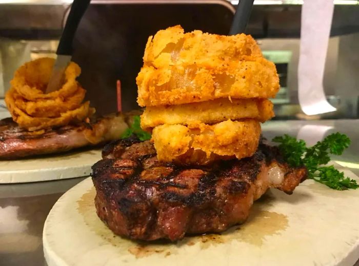 A large cut of meat garnished with a towering stack of onion rings, served on a plate alongside another order.