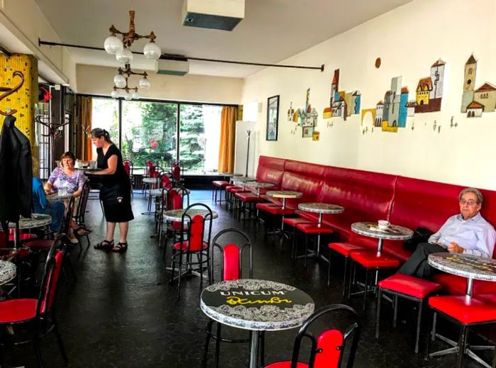 The cafe's interior features a mostly empty space with a few customers, a long red banquette lining one wall, and large windows at the back.