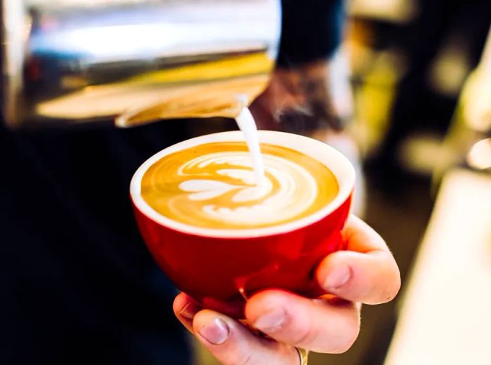 A barista skillfully pours milk into a red mug, creating a floral design in the coffee.