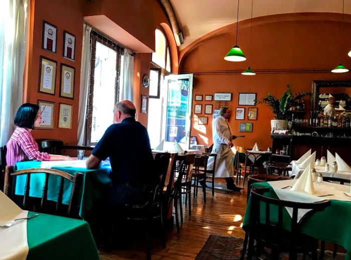 A cafe setting with a few diners at a table as someone enters. The walls are adorned with photographs, and the tables are dressed with green tablecloths.