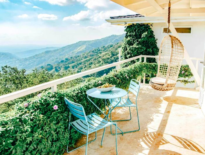 A terrace features a table with two chairs and a hanging swing, offering views over verdant hillsides and lush jungle.