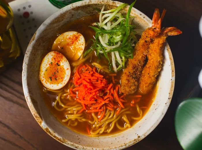 Aerial view of a stone bowl filled with ramen broth, topped with boiled eggs, scallions, carrots, and shrimp tempura.