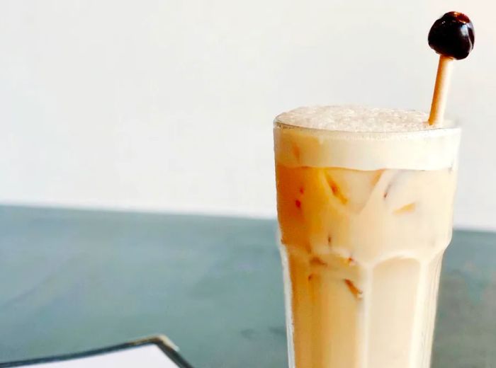 A tall, frothy cocktail garnished with a cherry on a long skewer, positioned next to a menu on a countertop.