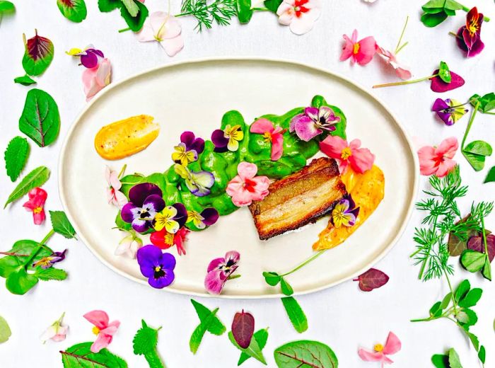 An aerial view of a serving platter featuring a large piece of cooked meat beside a green sauce smear, adorned with edible flowers, surrounded by flowers and leaves on a white background.