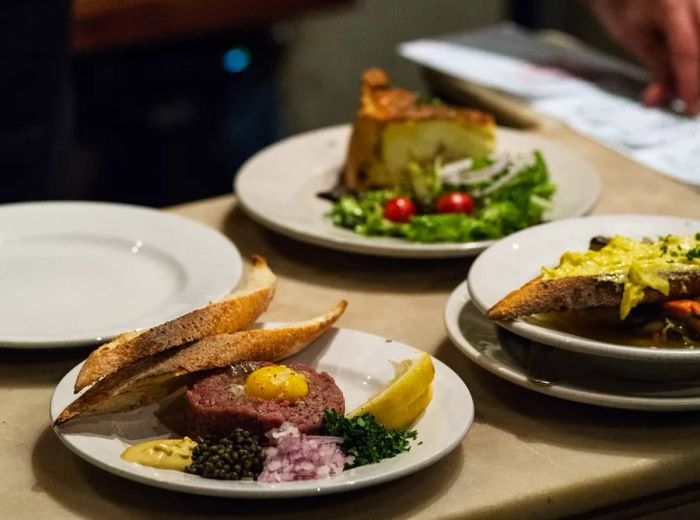 A countertop displaying a plate of egg-topped beef tartare, toast with rich yellow spread, and quiche accompanied by a salad.