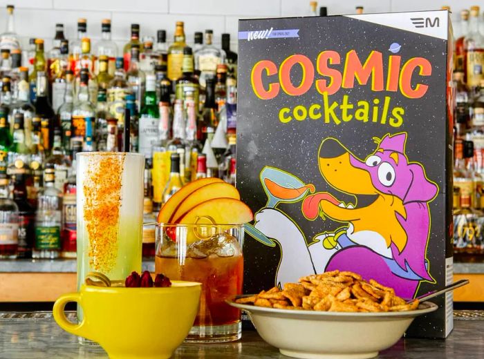 A whimsically branded cereal box beside a bowl of cereal and an array of cocktails on a bar top.