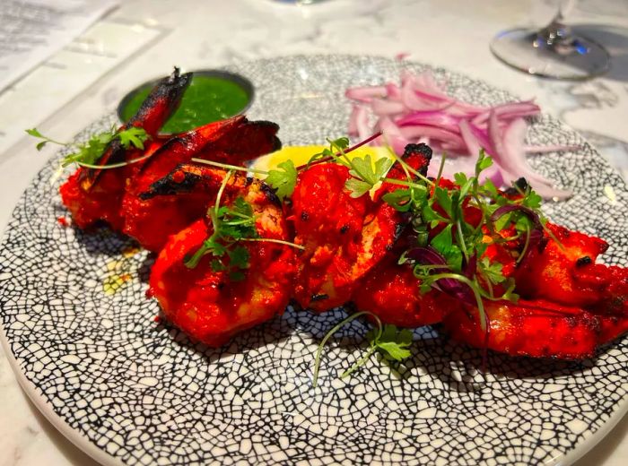 A few vibrant red shrimp arranged on a decorative plate, garnished with cilantro, sliced red onions, and a green dipping sauce.