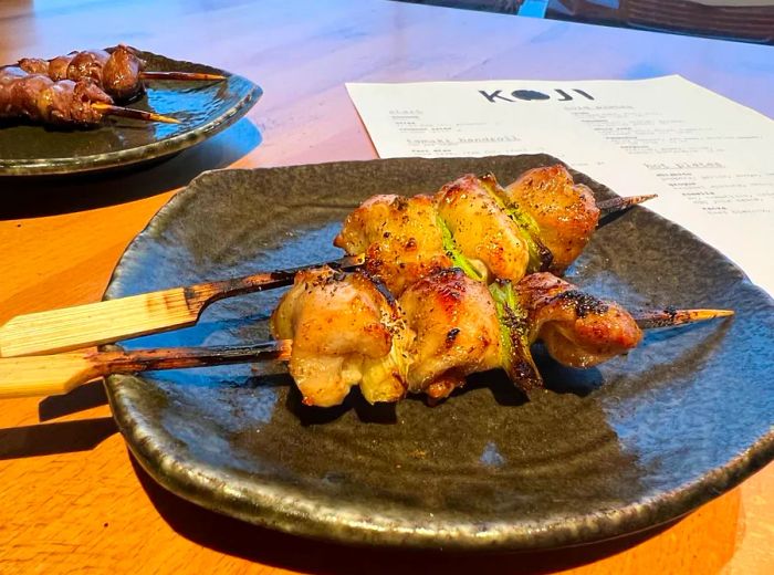 Two skewers of chicken, accompanied by additional skewers and a menu in the background.